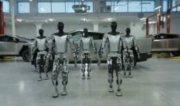 Vários robôs humanóides Tesla andando com alguns Cybertrucks ao fundo