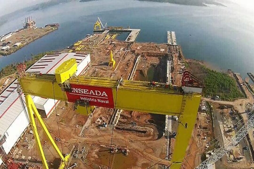 Vista aérea do Estaleiro Enseada em operação na Bahia, com guindastes e estruturas industriais à beira-mar.