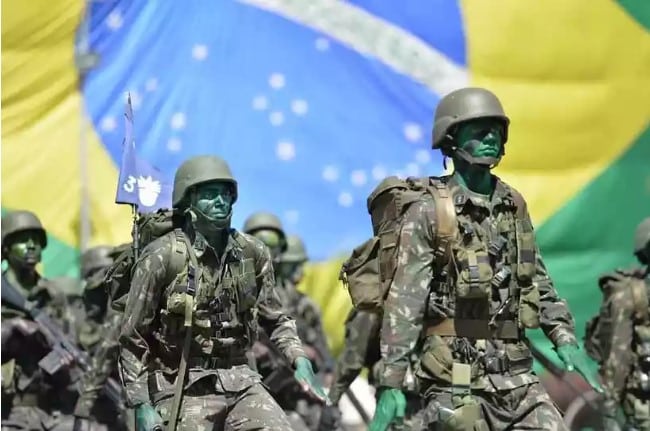 Tropa de sargentos do Exército Brasileiro marchando com uma bandeira do Brasil hasteada ao fundo