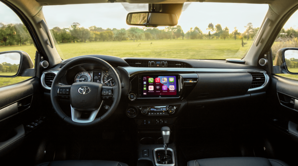 O painel da Toyota Hilux SRV 2025 conta com velocímetro e conta-giros analógicos, além de uma pequena tela digital para informações como consumo, autonomia e ajustes do veículo. A central multimídia de 8 polegadas tem Android Auto e Apple CarPlay sem fio, enquanto o ar-condicionado é digital dual zone; há ainda volante multifuncional revestido em couro e retrovisor interno eletrocrômico.