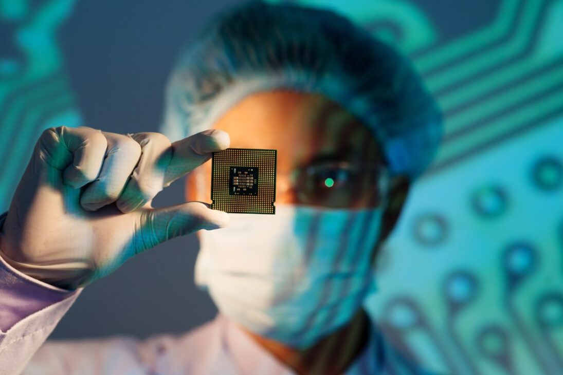 Cientista com máscara, touca e luvas segurando um microchip, representando pesquisa, inovação e alta tecnologia em eletrônica.

