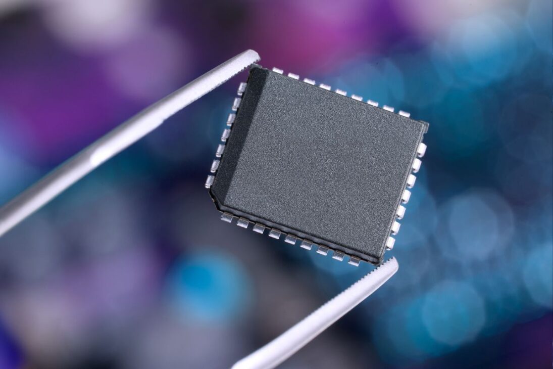 Microchip eletrônico sendo cuidadosamente segurado por uma pinça, com fundo desfocado em tons azulados, representando precisão na fabricação de componentes de alta tecnologia.

