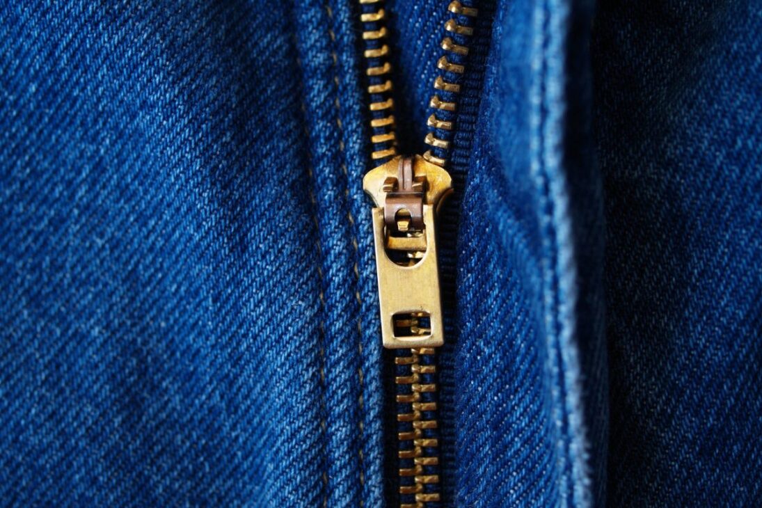 Zíper de metal dourado em uma calça jeans azul, com costura contrastante e a textura do tecido denim em destaque.