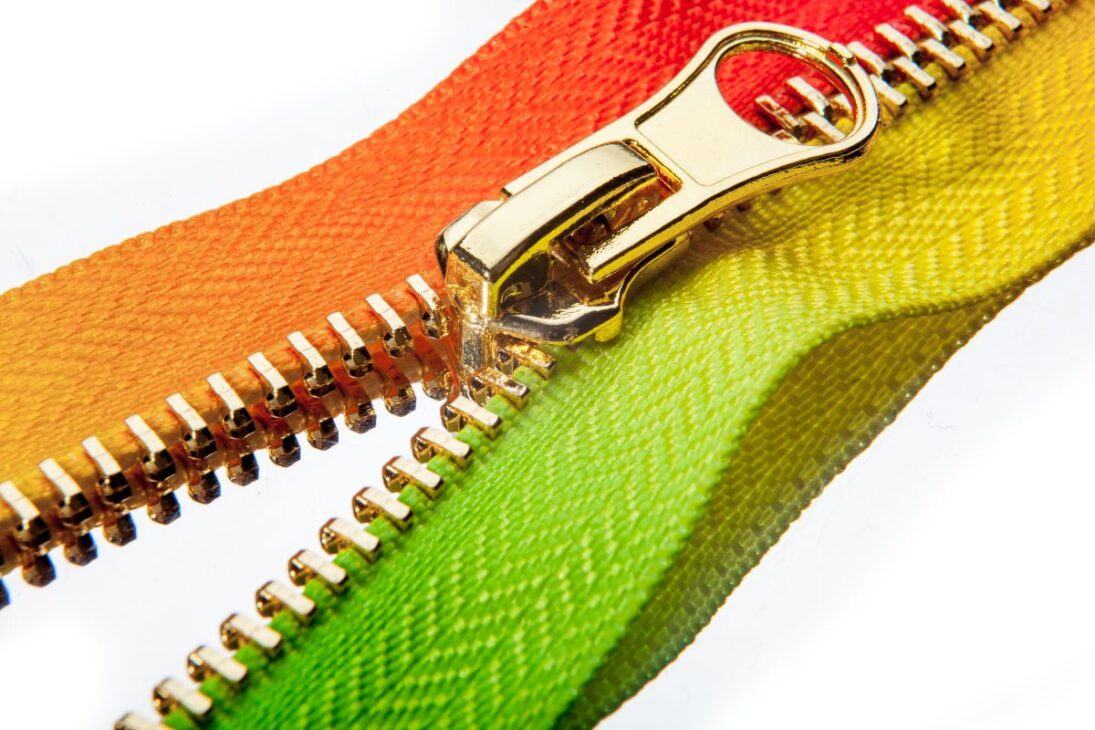 Zíper de metal dourado sobre um fundo branco, conectando tecidos com cores vibrantes: laranja, verde-limão e amarelo, mostrando a funcionalidade do fecho.
