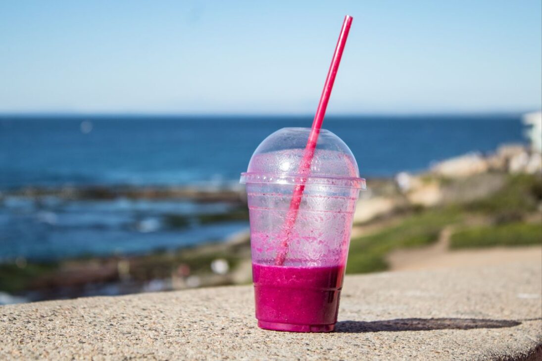 Um copo de plástico quase vazio com restos de bebida roxa e um canudo rosa, em um muro com o oceano ao fundo.