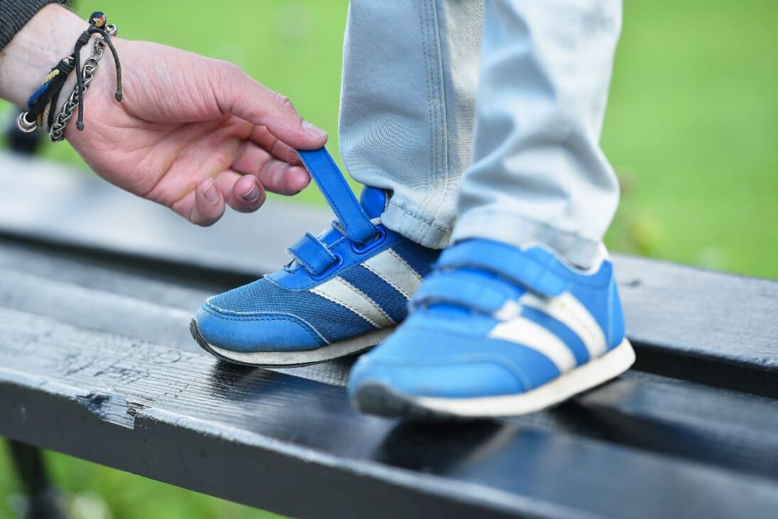 Pés de uma criança com tênis azul e branco com fechos de velcro, com a mão de um adulto ajustando uma das tiras.