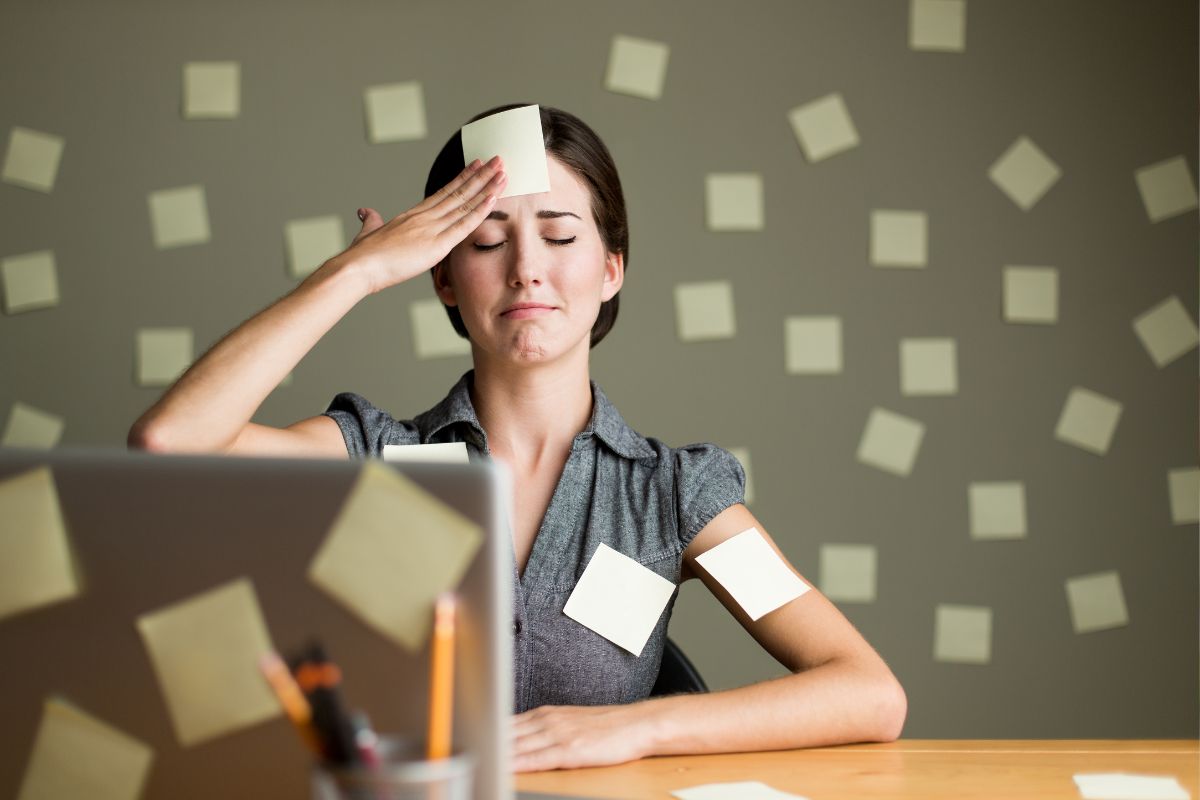 Gestresste Frau mit Post-its im Gesicht und am Arm, sitzt vor einem Laptop.