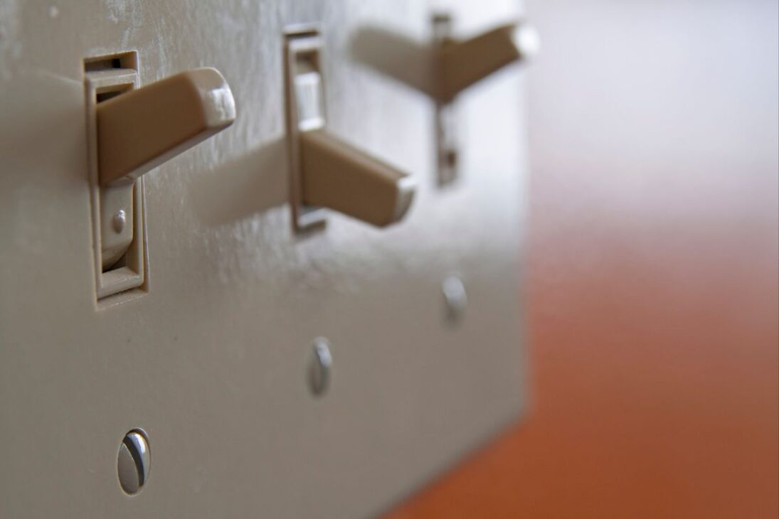 Três interruptores de luz bege, estilo alavanca, em uma placa de parede, com os dois primeiros na posição "ligado" e o terceiro parcialmente visível ao fundo.