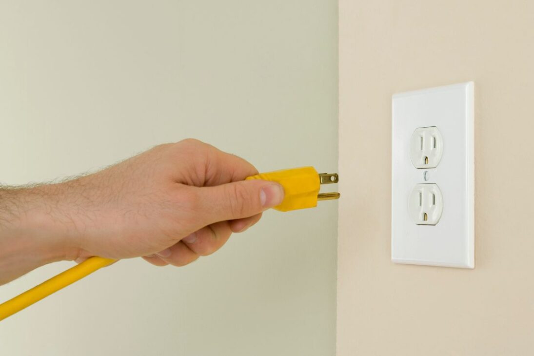 Mão inserindo um plugue amarelo de três pinos (Tipo B) em uma tomada elétrica americana branca de dois soquetes.