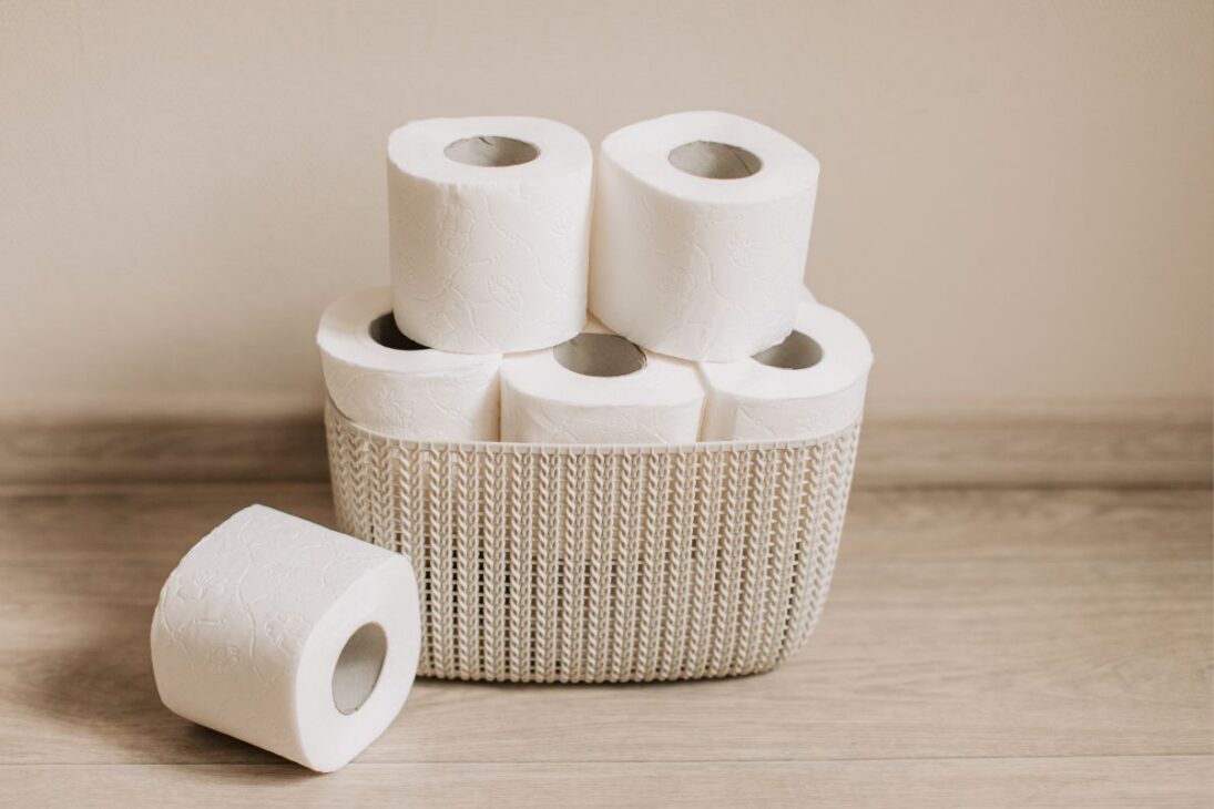 Cesto de vime com rolos de papel higiênico branco empilhados em um piso de madeira clara.