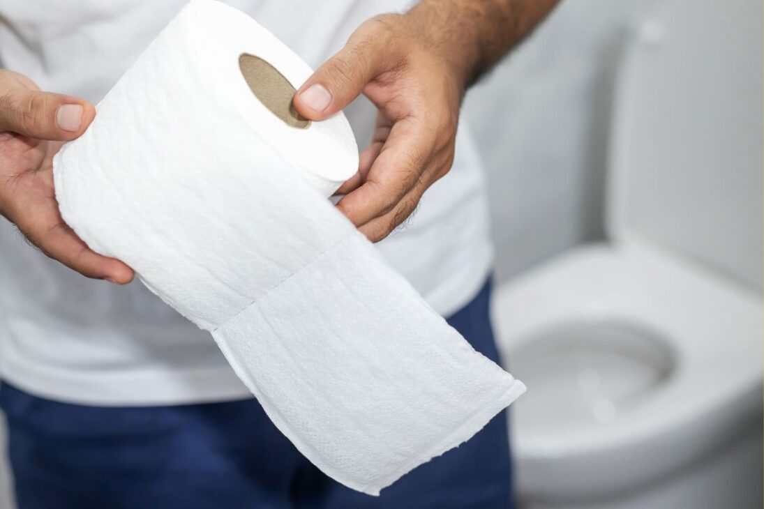 Mãos segurando um rolo de papel higiênico com um vaso sanitário desfocado ao fundo.