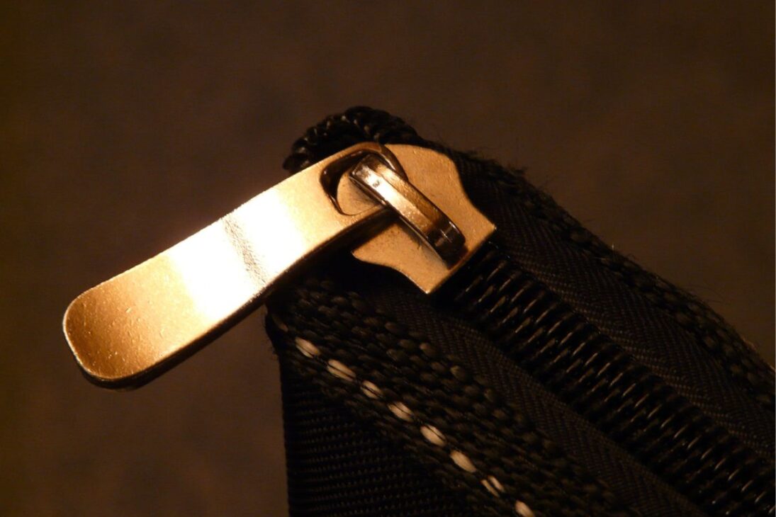 Zíper metálico dourado em close-up, preso a um tecido preto texturizado, com iluminação quente destacando o brilho do metal.