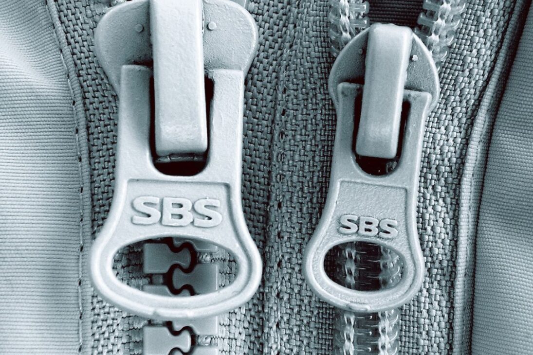  Dois puxadores de zíper cinzentos, marcados "SBS", em tamanhos ligeiramente diferentes, sobre um tecido texturizado de cor clara.