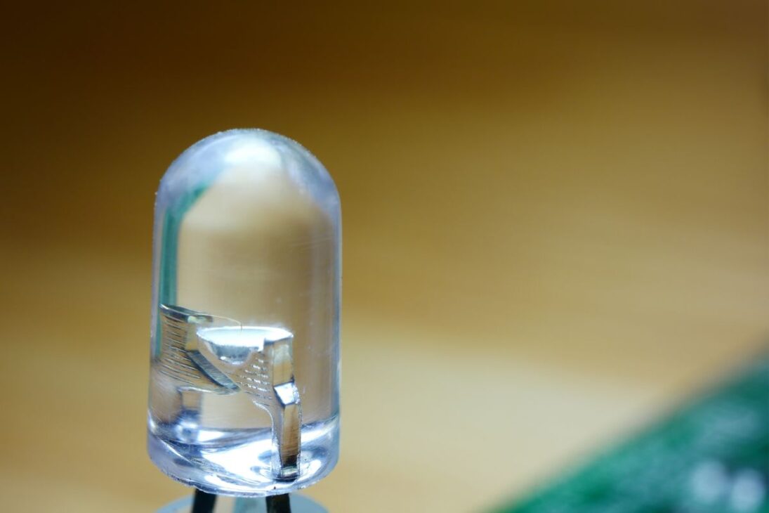 Um diodo emissor de luz (LED) transparente com foco nos componentes internos, como o chip semicondutor e o terminal de chumbo, contra um fundo desfocado de madeira e circuito verde.