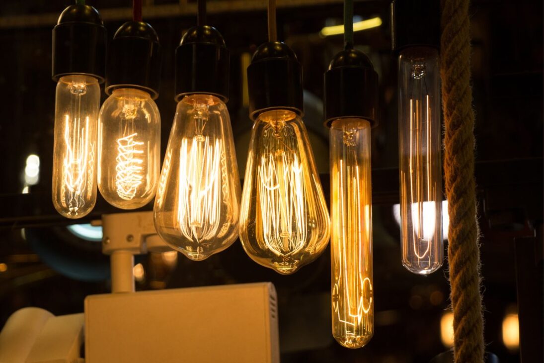 Seis lâmpadas de filamento estilo Edison penduradas, emitindo um brilho quente e laranja, contrastando com um fundo escuro.