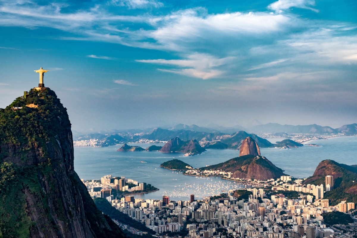 Vista aérea do Rio de Janeiro com o Cristo Redentor no Morro do Corcovado à esquerda e o Pão de Açúcar ao fundo, com a Baía de Guanabara e prédios ao redor.