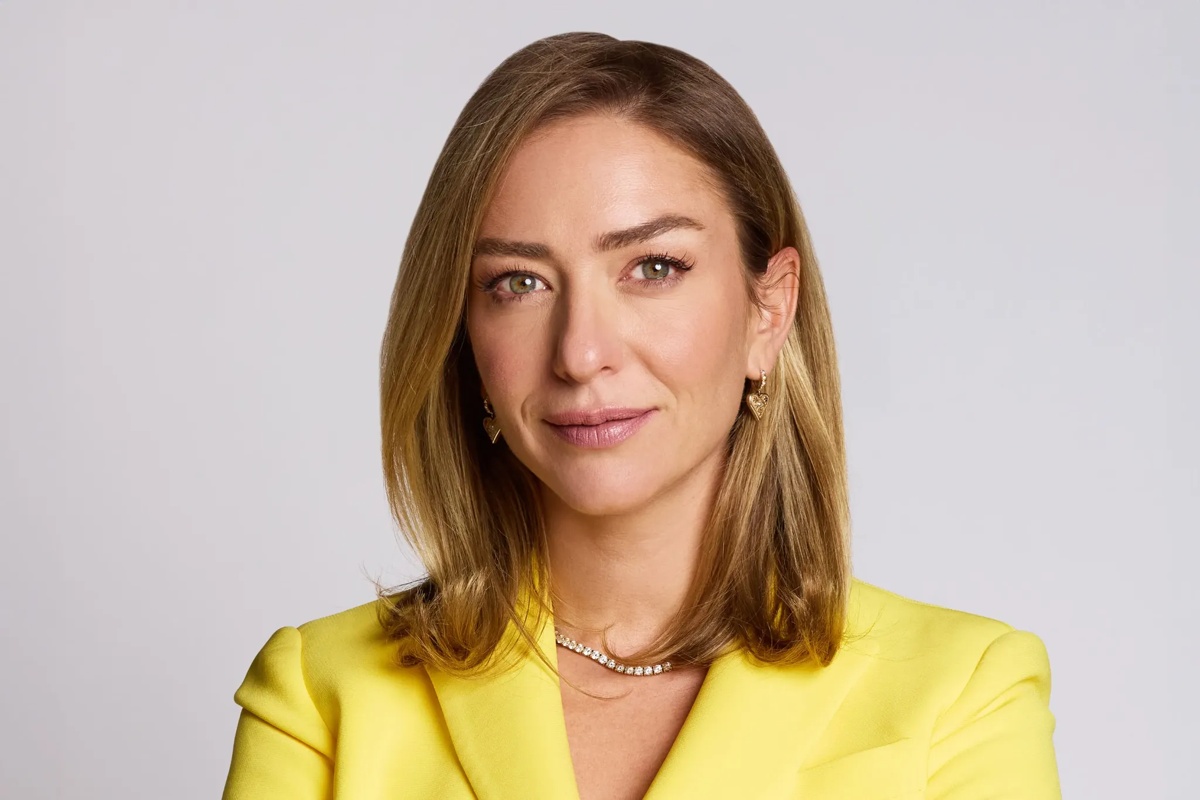 Retrato de uma mulher de cabelo loiro, usando blazer amarelo e joias douradas, com expressão confiante em fundo claro.