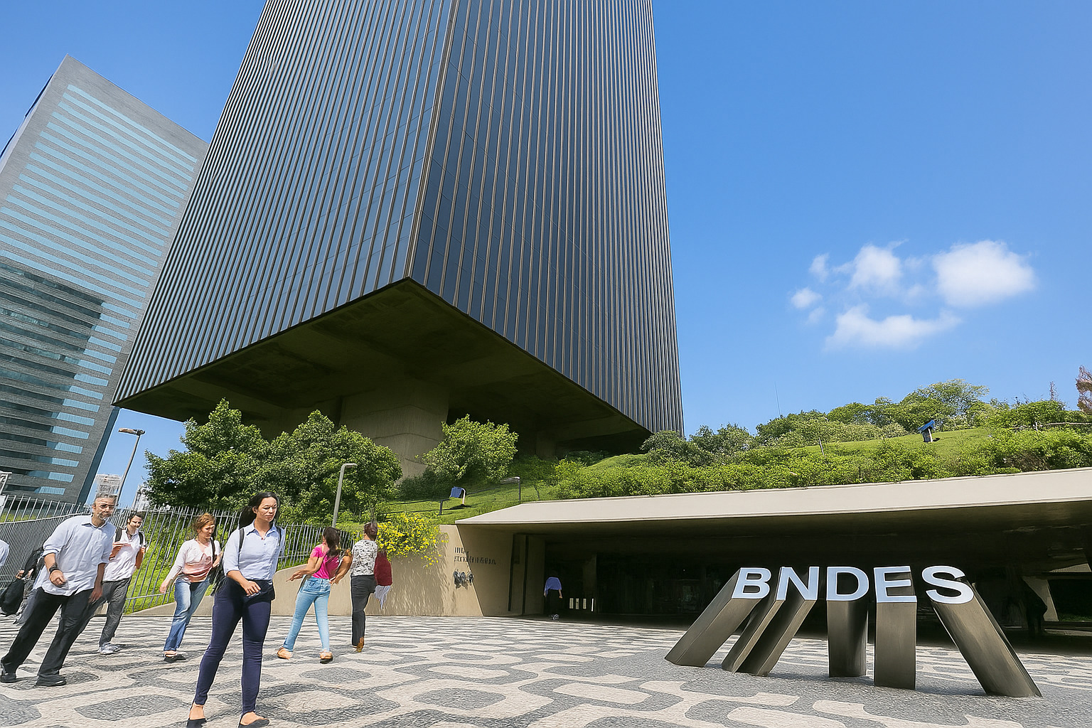Fachada do edifício do BNDES em dia claro com céu azul e poucas nuvens, com pessoas caminhando na entrada principal.