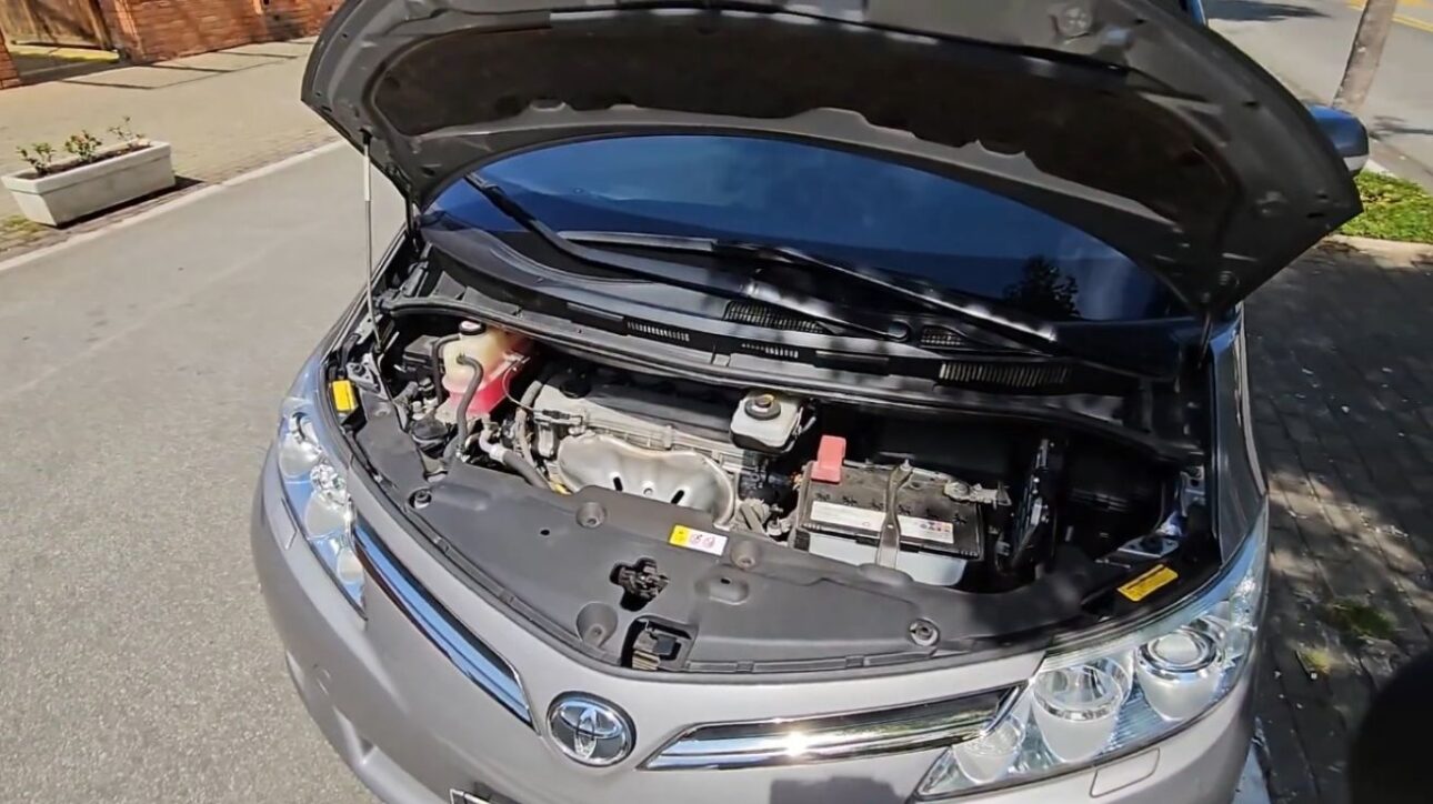 Compartimento do motor da Toyota Previa 2018 com o capô aberto, exibindo o motor 2.4 a gasolina e componentes mecânicos, em rua urbana durante o dia.