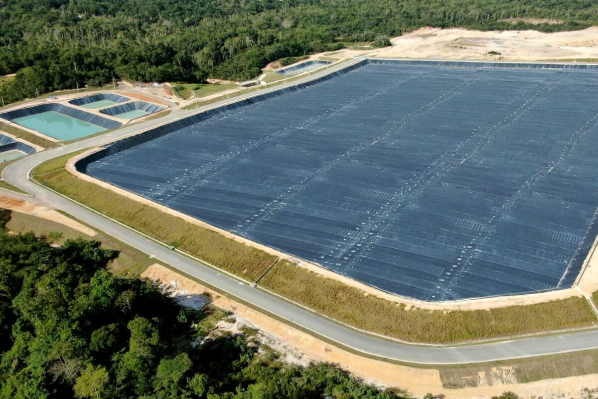 Vista aérea de um aterro sanitário com grandes áreas cobertas por lonas pretas e tanques de contenção de líquidos ao lado, rodeado por vegetação e estrada pavimentada.