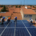 Profissionais instalando painéis solares em telhado residencial com céu azul ao fundo.