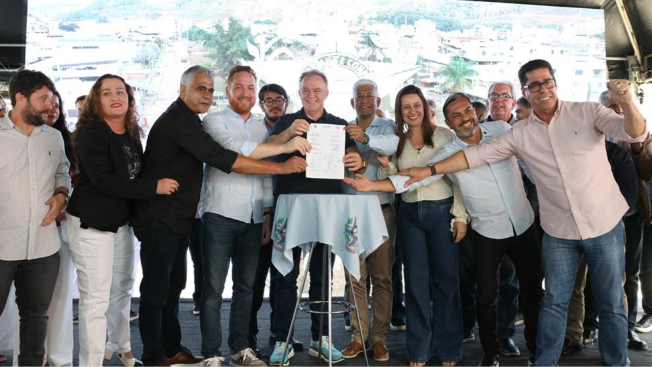 Grupo reunido em cerimônia pública ao ar livre, com destaque para documento apresentado por figura central.
