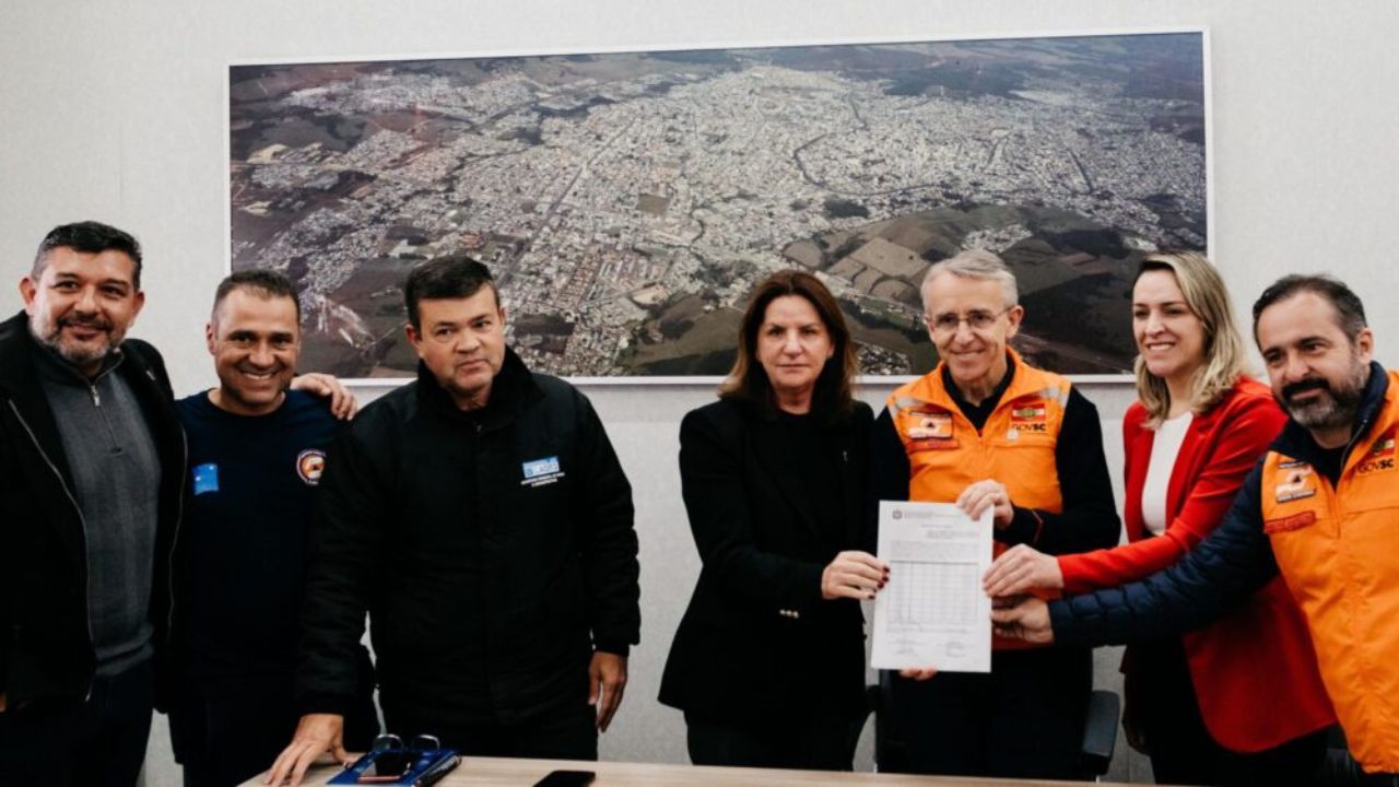 Grupo de autoridades e técnicos da Defesa Civil de SC posa com documento oficial em frente a imagem aérea de Lages, simbolizando o anúncio de obras de prevenção e infraestrutura urbana.