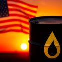 Barril de petróleo preto com símbolo dourado em destaque, ao pôr do sol, com a bandeira dos Estados Unidos ao fundo em um campo aberto