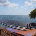Trabalhadores instalam painéis solares flutuantes sobre a água em um lago sob céu parcialmente nublado.