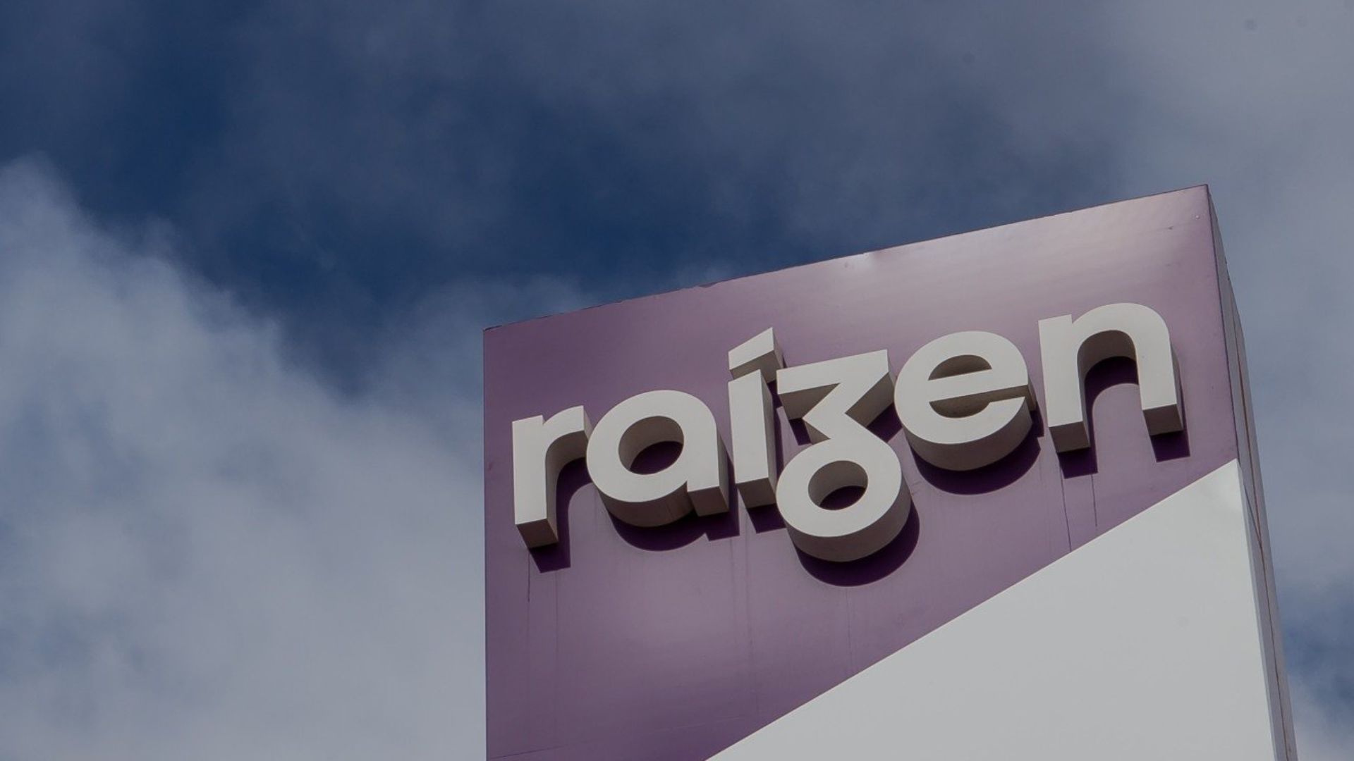 Totem vertical da Raízen com logotipo branco sobre fundo roxo, fotografado contra o céu nublado.