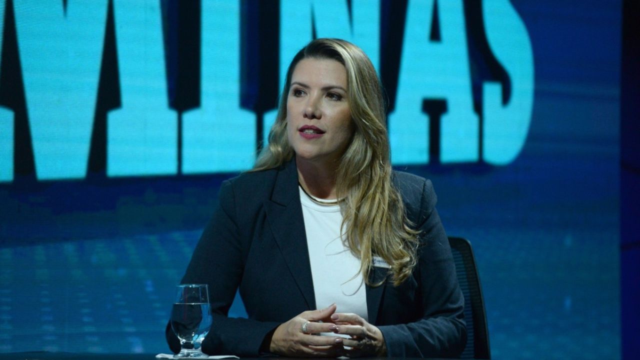Prefeita de Uberaba vestindo blazer escuro e blusa branca, sentada em mesa de estúdio com fundo azul e letras grandes ao fundo, em ambiente de entrevista ou programa televisivo.