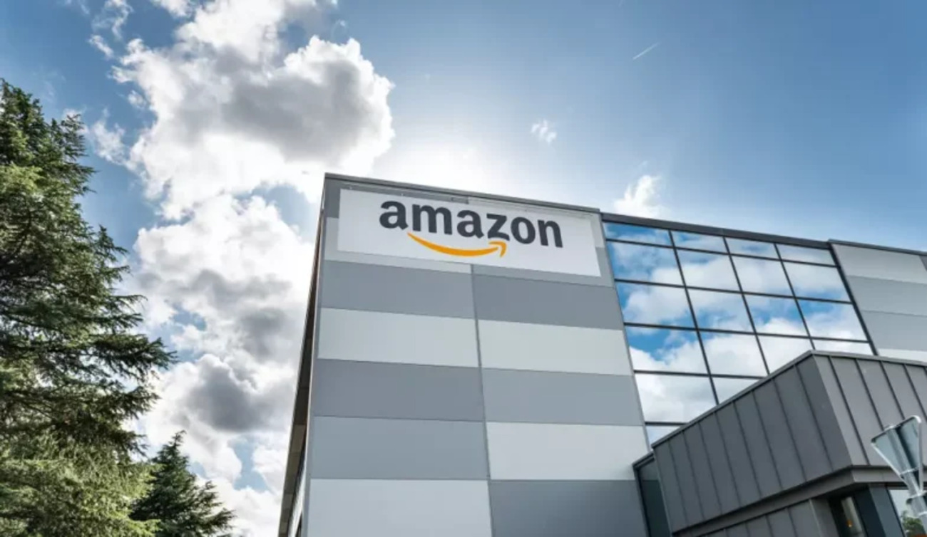 Fachada de prédio corporativo da Amazon com logotipo da empresa em destaque sob céu azul com nuvens.
