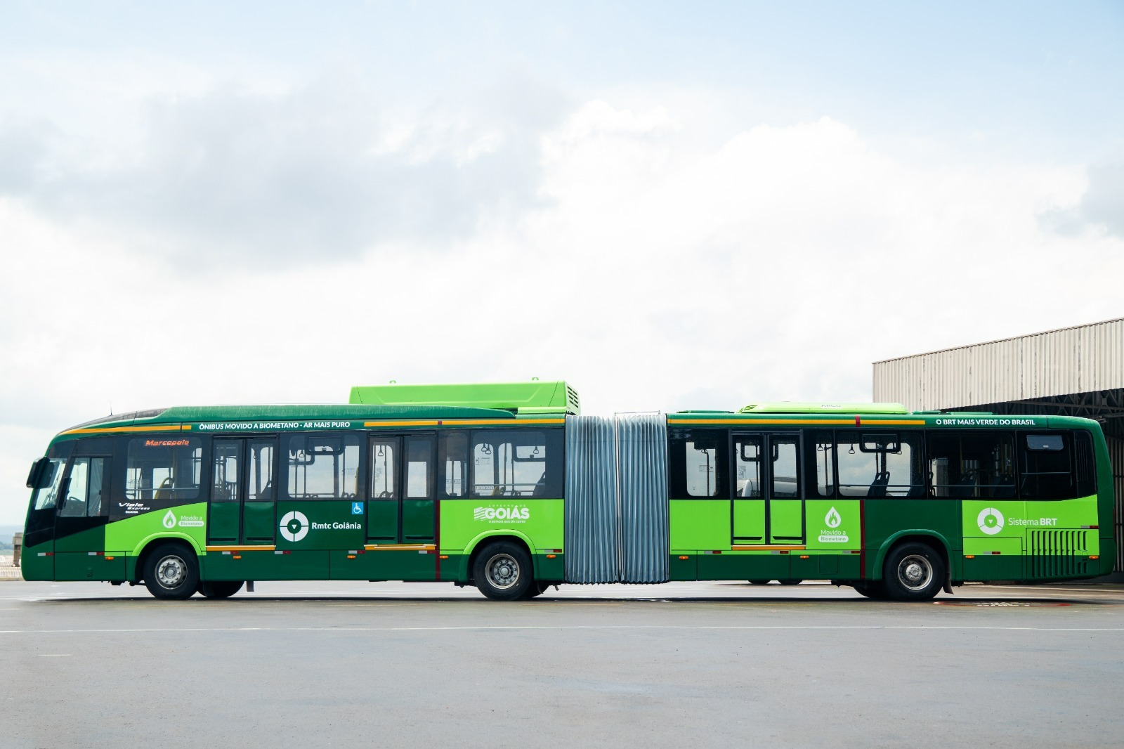 Ônibus articulado verde movido a biometano estacionado em área urbana, com design moderno e foco em transporte sustentável.