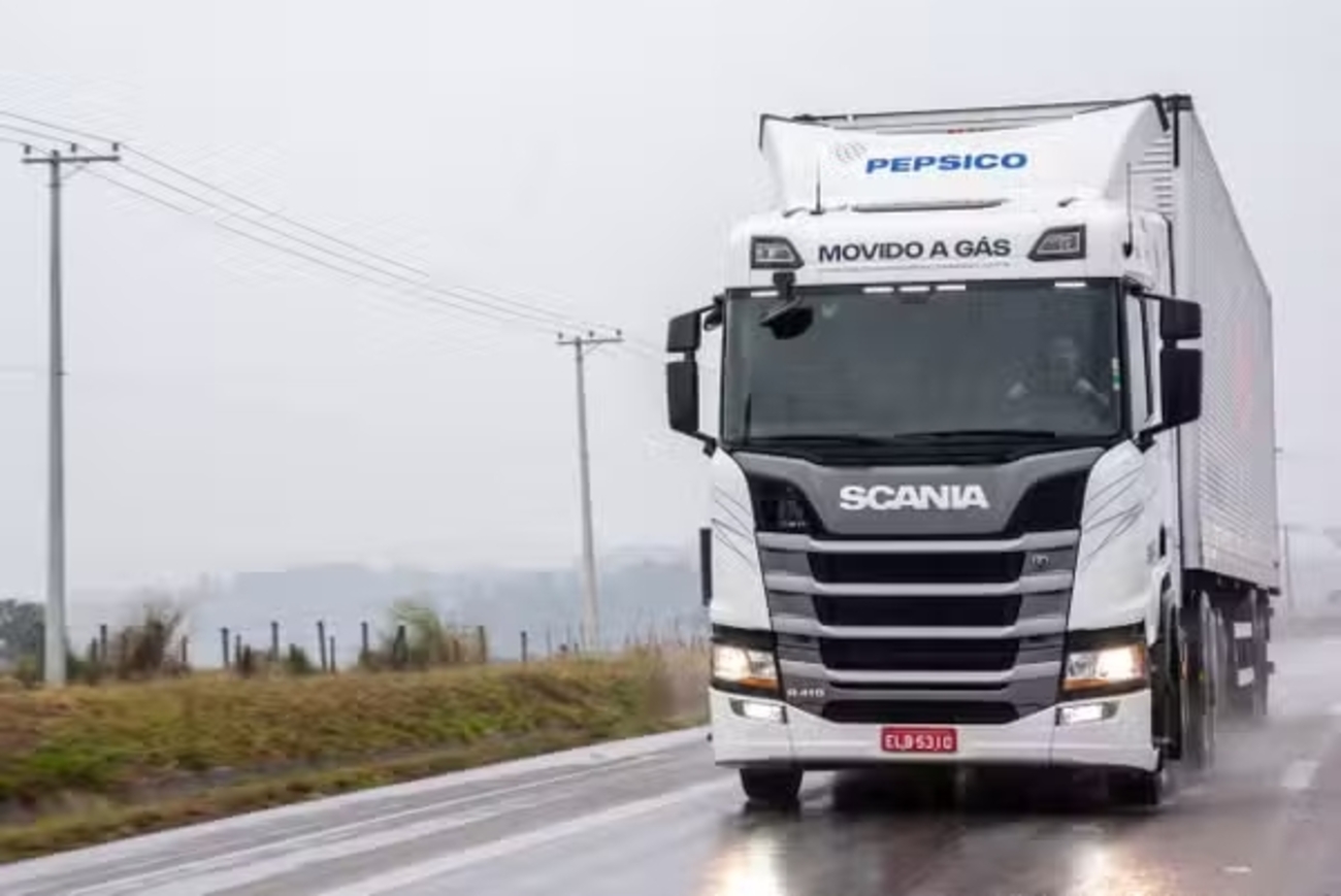 Caminhão Scania da PepsiCo movido a gás trafegando em estrada molhada em área rural.