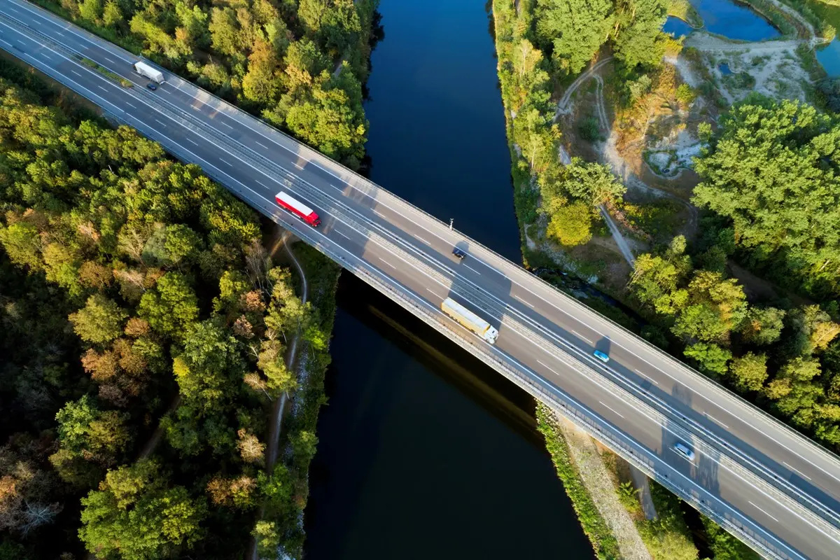 Vista aérea de rodovia com caminhões e carros atravessando ponte sobre rio cercado por vegetação.