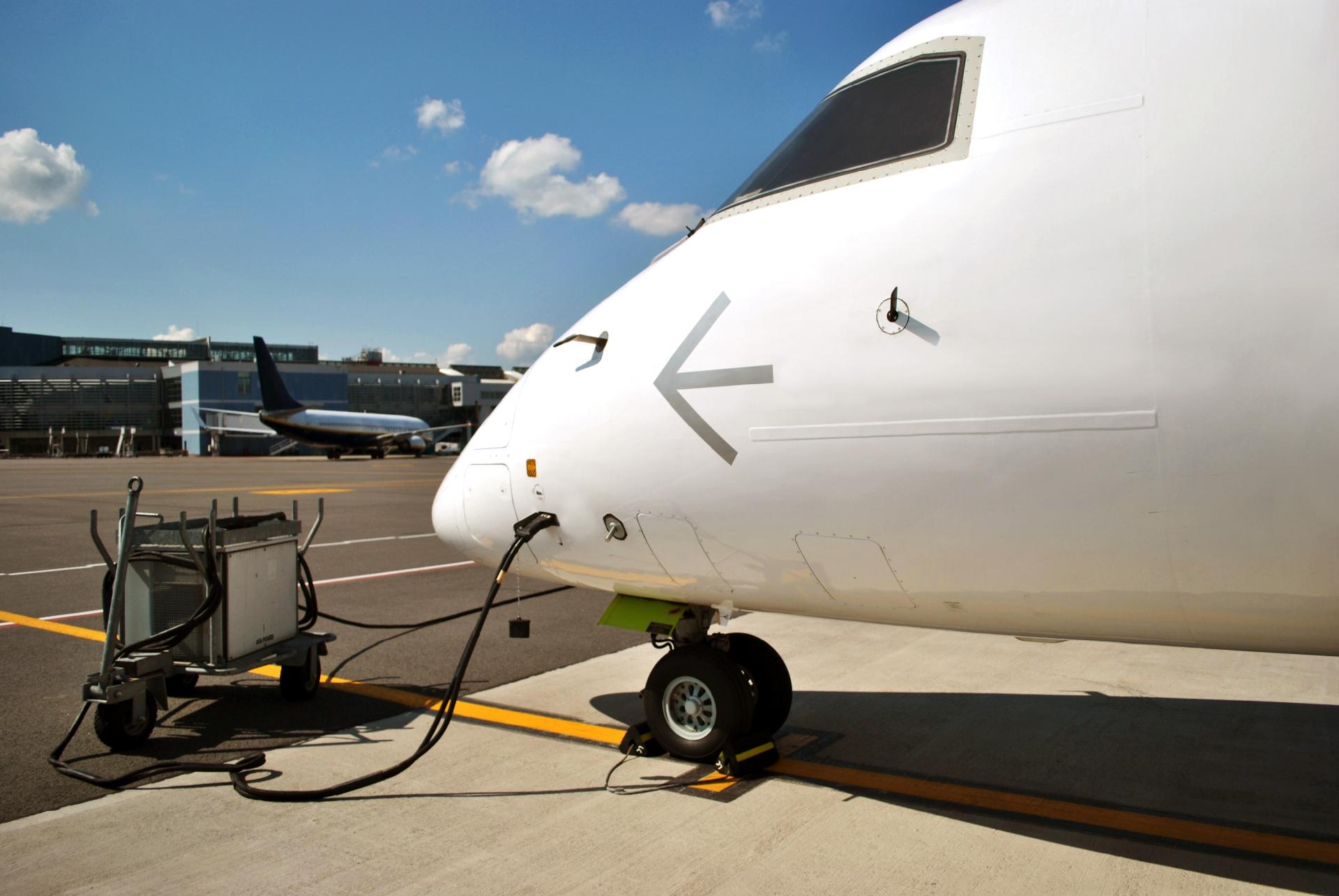 Avião estacionado no aeroporto sendo abastecido com combustível por equipamento conectado à fuselagem.