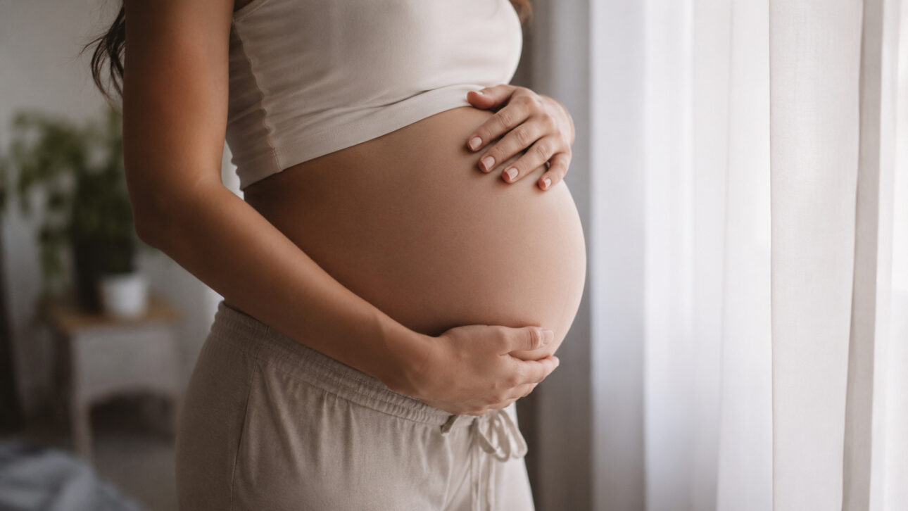 Gestante segura a la barriga cerca de una ventana iluminada, simbolizando planificación familiar, embarazo y la importancia de las pruebas genéticas para evaluación de enfermedades hereditarias antes de la concepción.