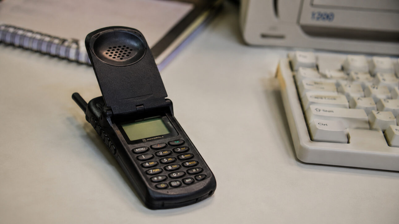 Foldable phone inspired by the Motorola StarTAC on an office table, next to a keyboard and old monitor, representing the mobile technology used in the early 2000s.