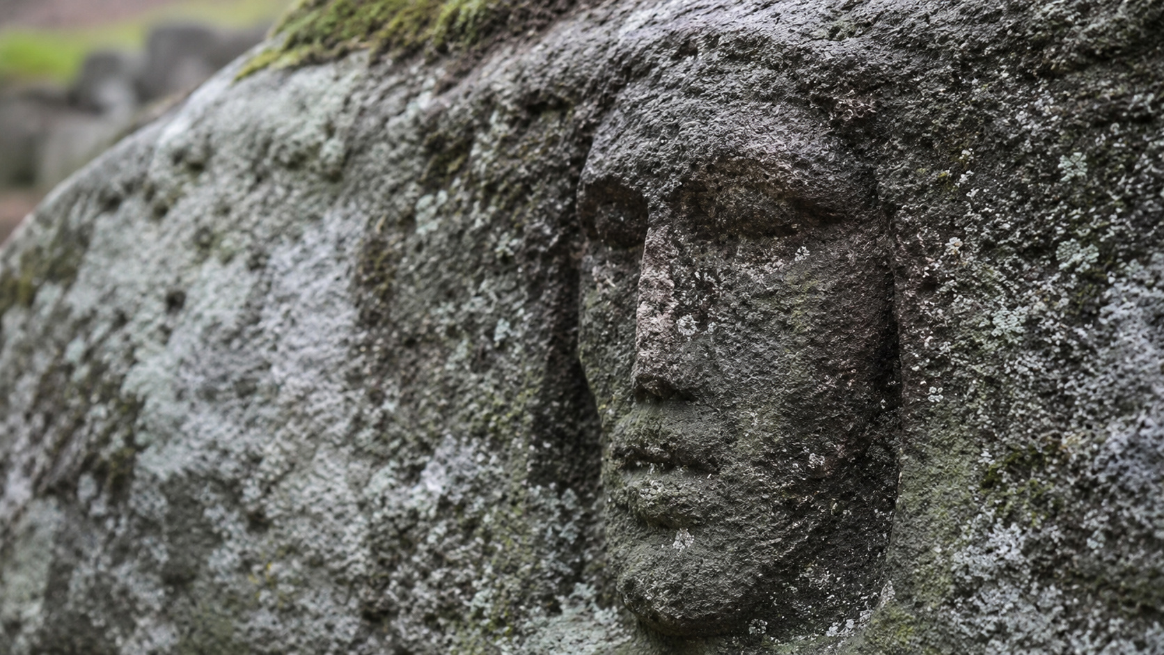 Rosto esculpido em pedra antiga, com textura desgastada e musgos, representando artefato arqueológico ligado a rituais funerários da Idade do Bronze no Cazaquistão.