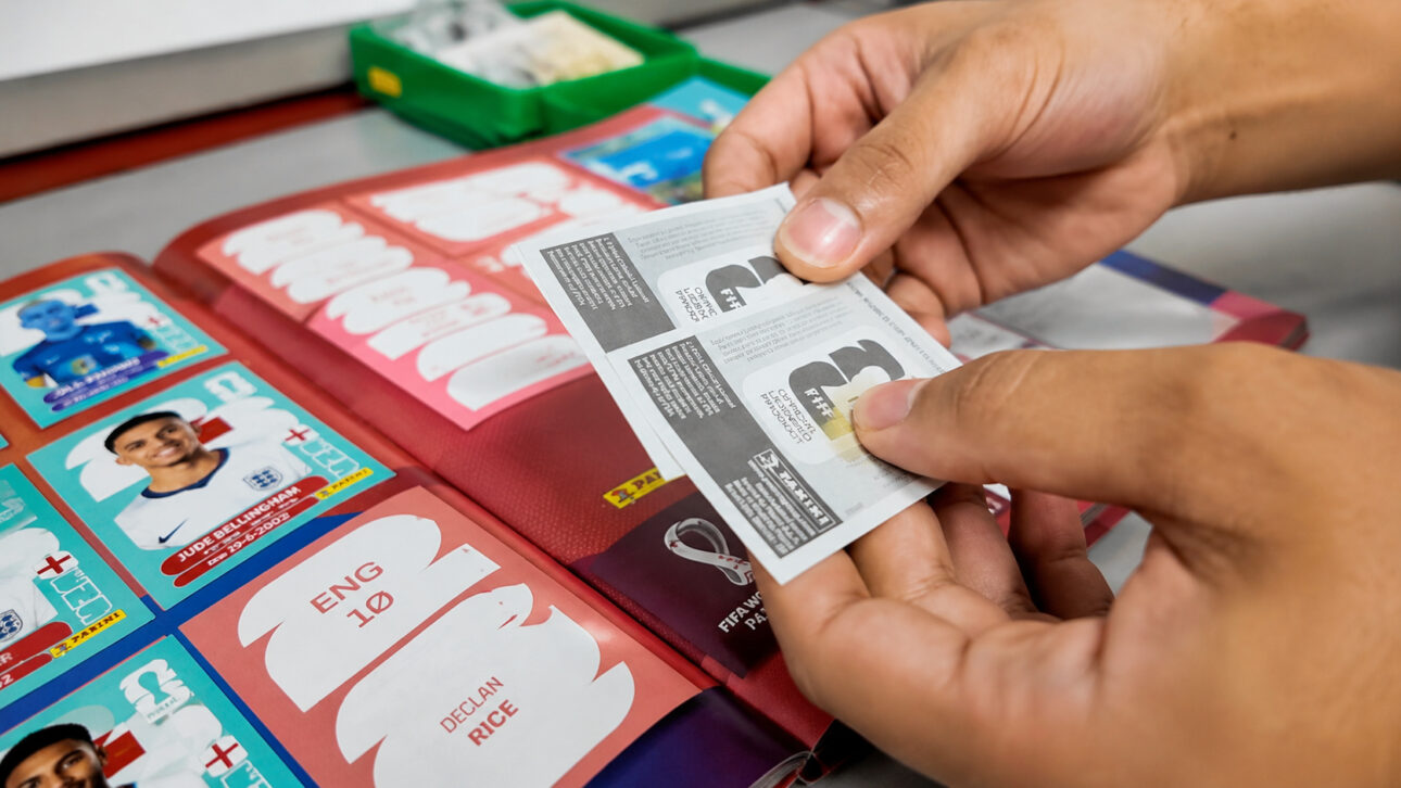 Hands holding the discarded protective paper from World Cup stickers over an open album, highlighting the waste used in a recycling project developed by a Brazilian student.