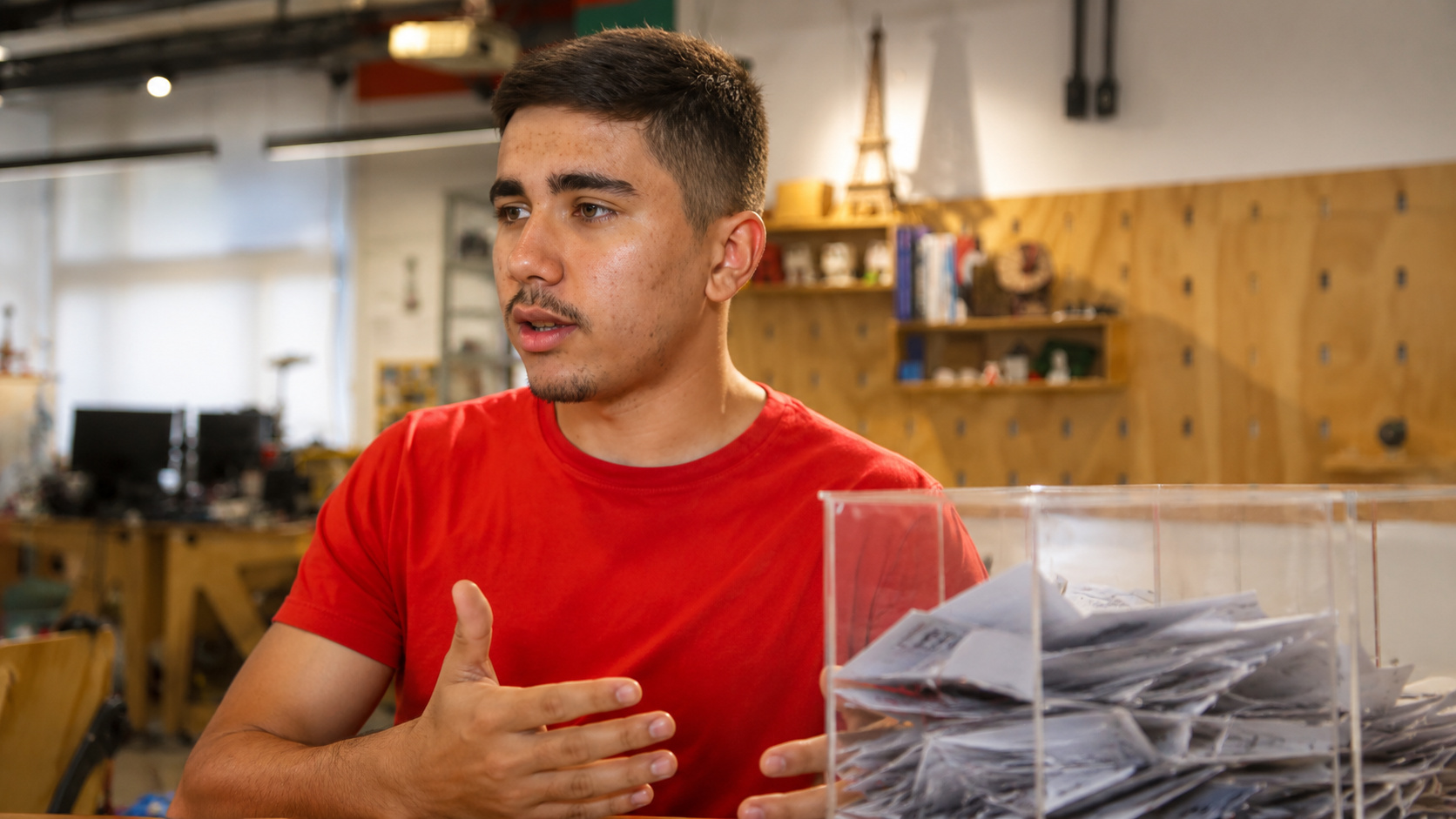 Estudante responsável pelo projeto de reciclagem de papéis de figurinhas da Copa apresenta a iniciativa ao lado de uma urna de coleta em ambiente educacional e tecnológico.