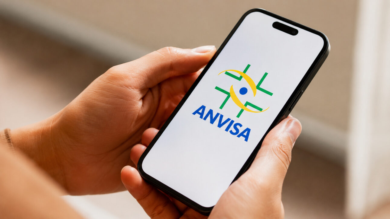 Pessoa segura smartphone com a identificação da Anvisa em matéria sobre lotes suspensos de creatina, BCAA, beta-alanina e multivitamínicos.