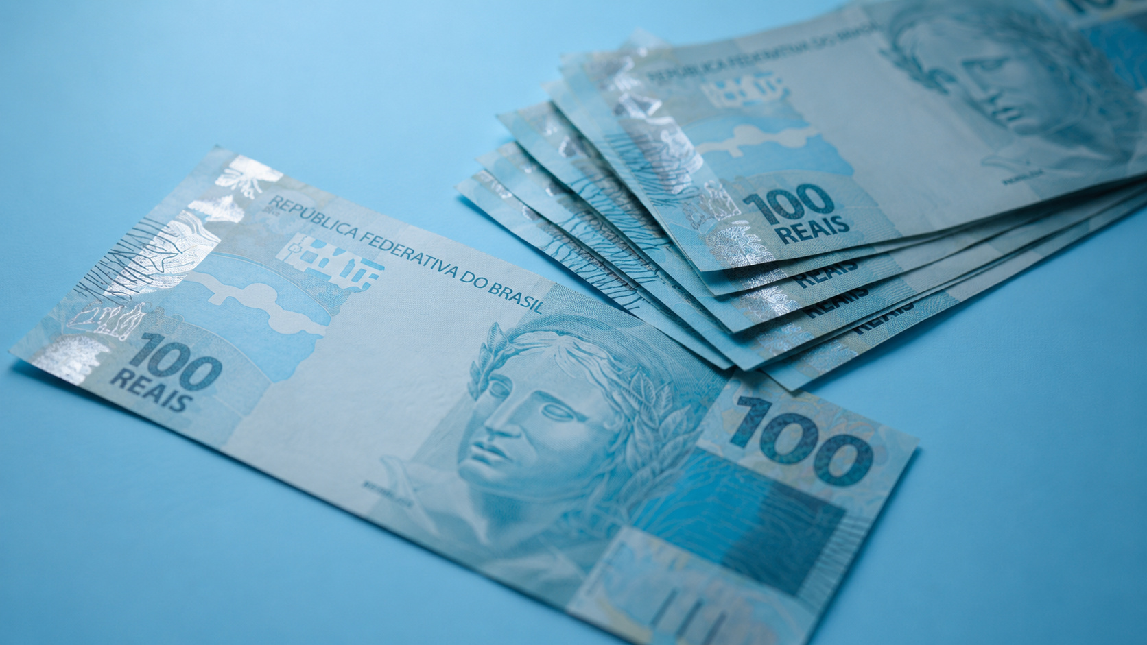 Cédulas de 100 reais em close sobre fundo azul, representando benefícios do CadÚnico, auxílio financeiro e redução de despesas para famílias de baixa renda.