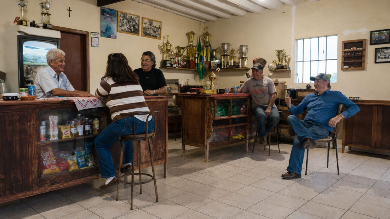Moradores reunidos em vendinha do interior de São Paulo com balcões de madeira, produtos, fotografias e troféus nas paredes.