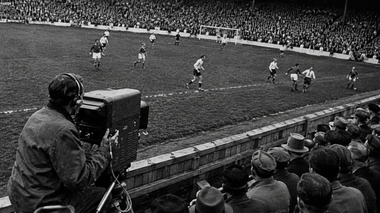 Cinegrafista com câmera antiga filma partida de futebol em estádio cheio, em imagem em preto e branco inspirada na Copa do Mundo de 1958.