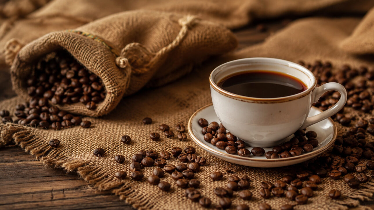 Xícara de café preto sobre tecido de juta, cercada por grãos torrados e um saco de café aberto.
