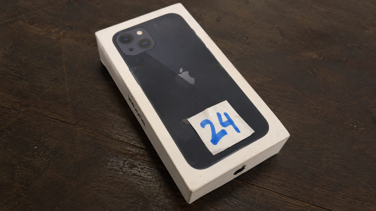 Caixa de iPhone 13 com etiqueta numérica sobre mesa de madeira, ilustrando lote do leilão da Receita Federal.