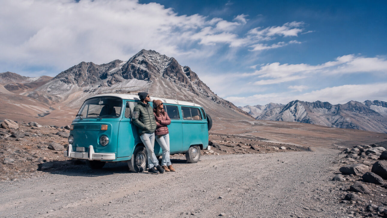 Casal de viajantes ao lado de uma Kombi azul em estrada rochosa, cercada por montanhas durante jornada pelas Américas.