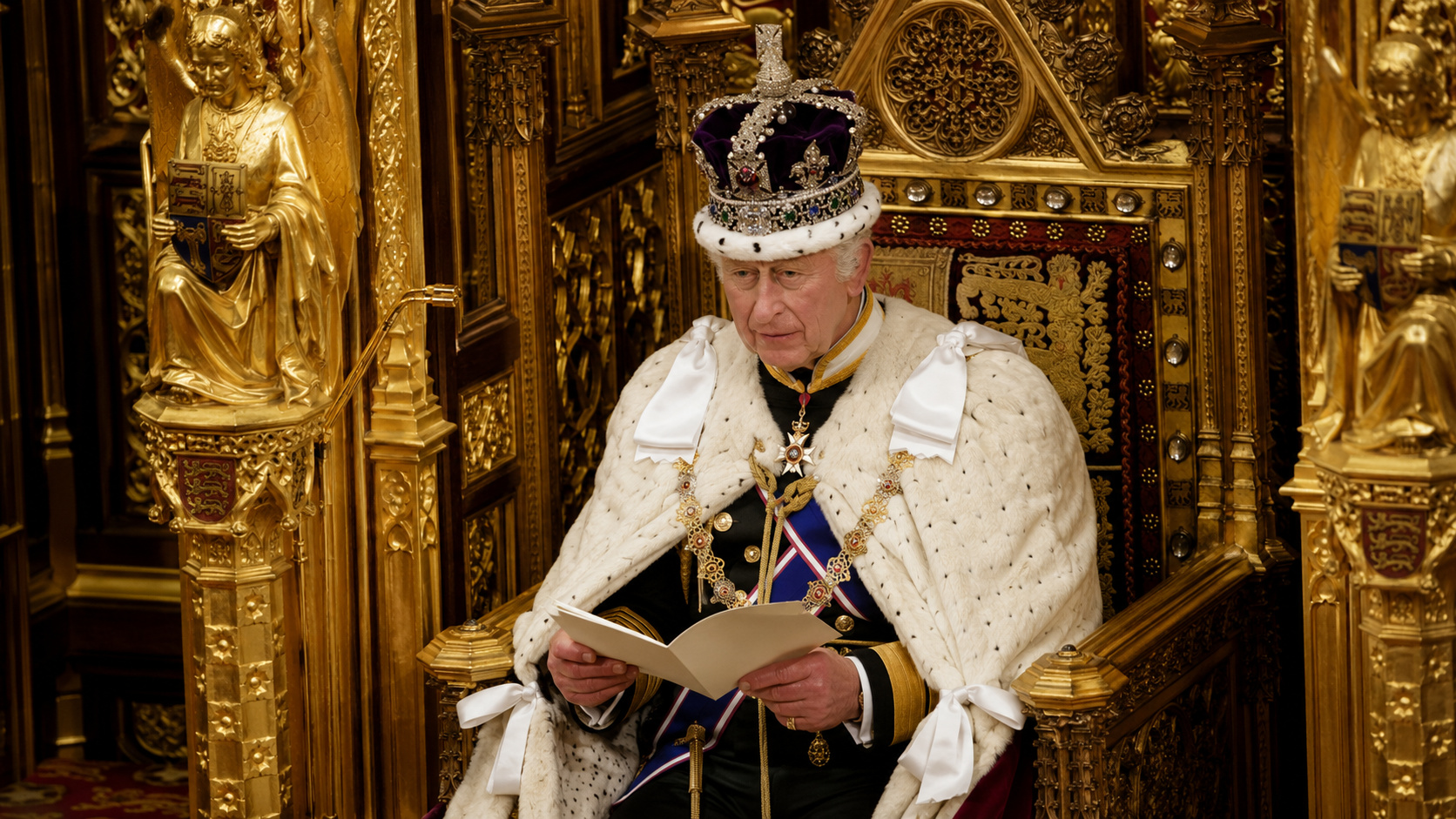 Rei Charles III com coroa e trajes cerimoniais sentado em trono dourado, segurando documento durante cerimônia da monarquia britânica.
