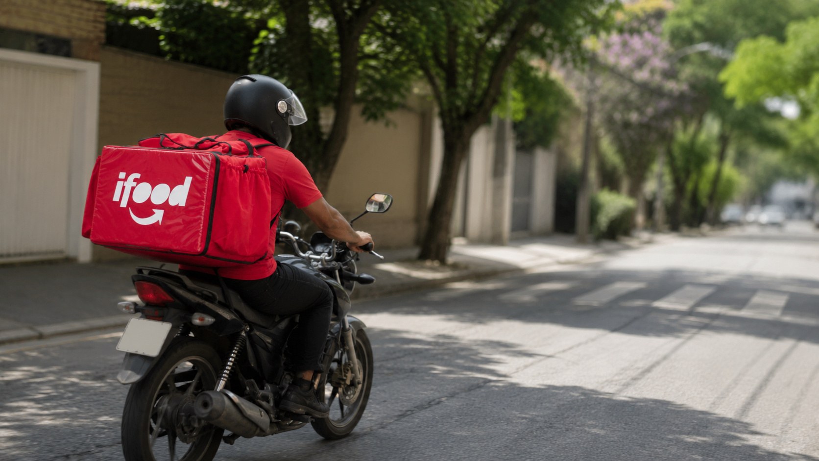 Entregador do iFood conduz motocicleta por rua arborizada durante serviço de delivery no Brasil